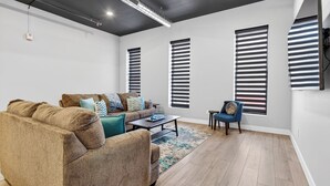 En moderne stue med sofa, sofabord og en stol. Tre vinduer med persienner, et loft med indbygningsbelysning og en synlig røgalarm.