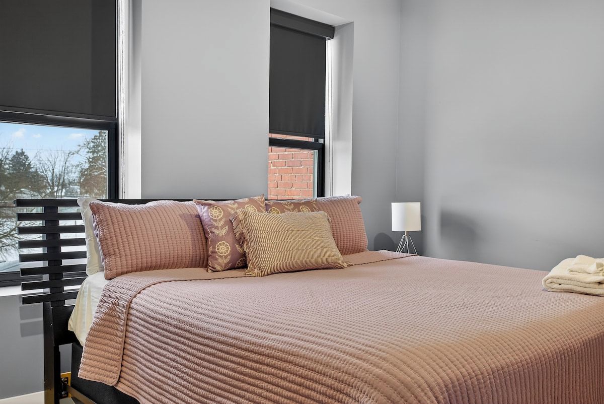 A bedroom with a bed, pillows, a nightstand, a lamp, and a window with blinds.