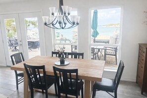 A dining area with a wooden table and black chairs, a chandelier, and a view of the sea through large windows.