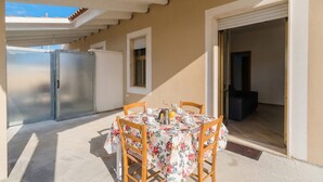Een terras buiten met een tafel, stoelen, een koelkast en een deur naar een binnenruimte.