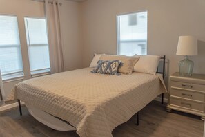 Un dormitorio con cama, mesita de noche, lámpara y dos ventanas con persianas.