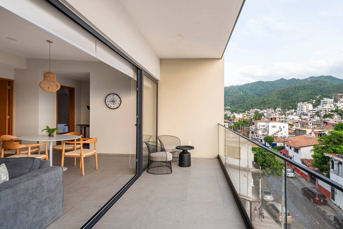 A balcony with a dining area, chairs, a sofa, a clock, and a view of a cityscape with mountains.