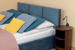 Ein Bett mit blauem Kopfteil, mehreren Kissen und einem Nachttisch mit Lampe.