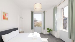 A hotel room with a large bed, a bedside table, a window with curtains, a radiator, and a potted plant. - Detached House with Sea Views & Parking (North Berwick)