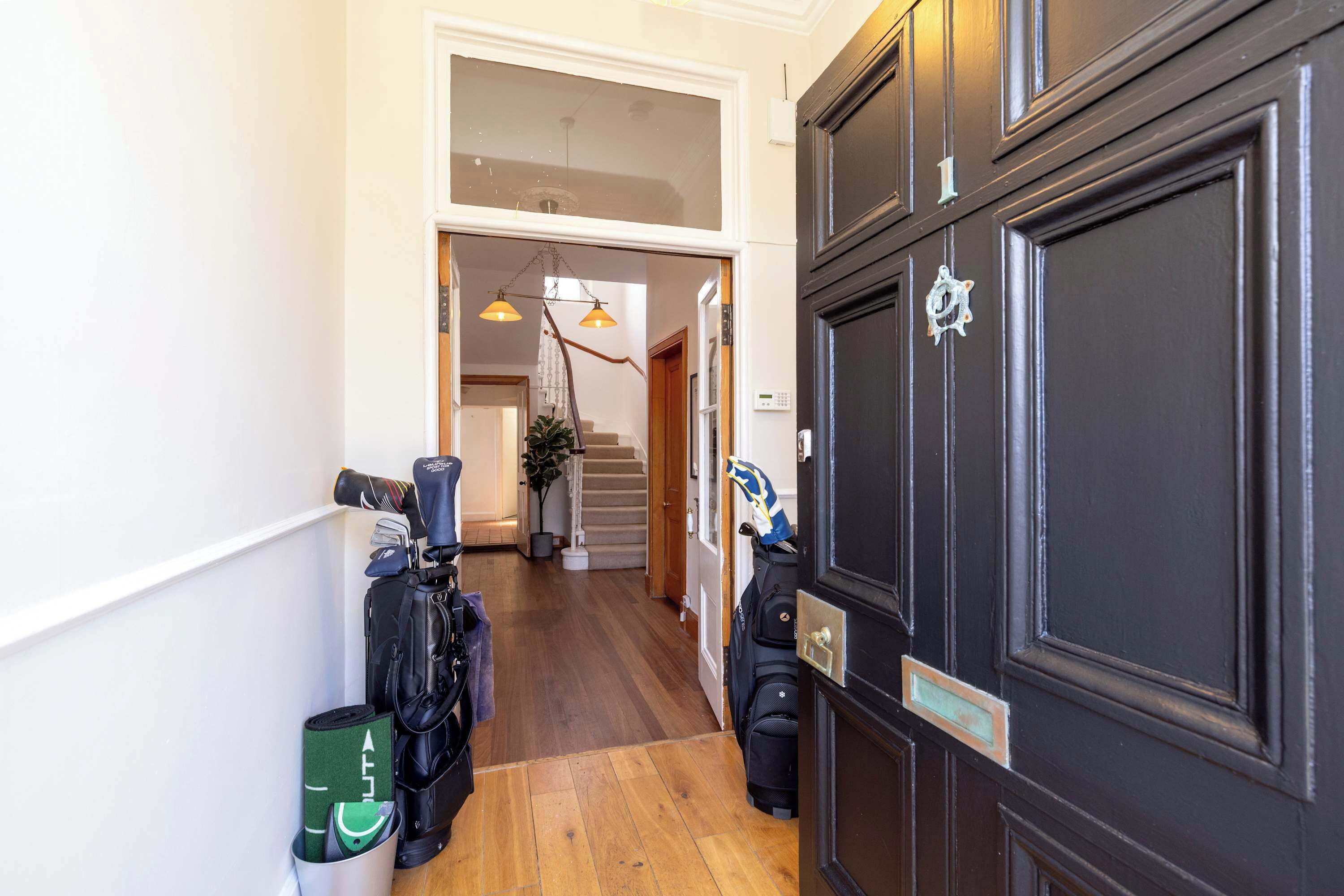 Un pasillo con piso de madera, una puerta negra con placa y una escalera que sube a un nivel superior.