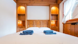 Una cama bien hecha con dos toallas dobladas, cabecera de madera con estantes integrados y lámparas fijadas a la pared.