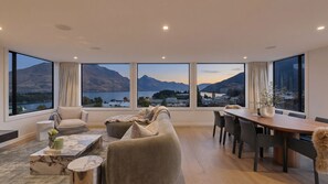 A modern living room with a large window offering a mountain and lake view, a sofa, a dining table, and a coffee table. - Queenstown Hill House Only -  A seamless sense of luxury for families or groups of friends. (Queenstown)