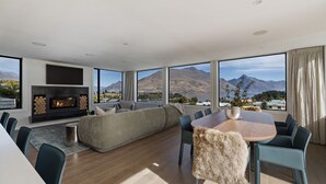 A modern living room with a fireplace, a large sofa, a dining table with chairs, and a view of mountains and a lake. - Queenstown Hill House Only -  A seamless sense of luxury for families or groups of friends. (Queenstown)