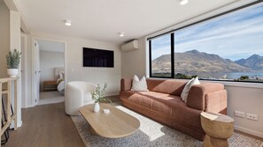 A modern living room with a sofa, coffee table, and a large window offering a mountain view. - Queenstown Hill House - Apartment Only with stunning lake & mountain views. (Queenstown)