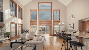 A spacious living area with a high ceiling, large windows, a stone fireplace, and a kitchen with a bar area. - 892 Saddle Ridge Ultra luxurious 4 bedroom home, views, 2 living spaces and more (Granby)