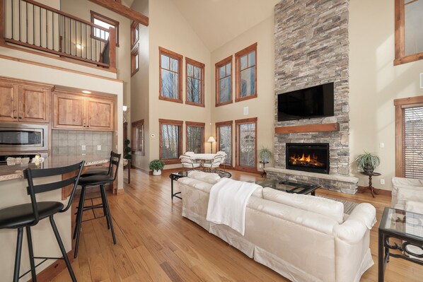 A spacious living area with a fireplace, wooden flooring, and a high ceiling.