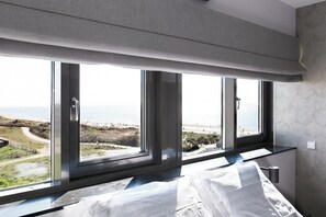 Ein Schlafzimmer mit großem Fenster mit Blick auf einen Strand, einem Bett und einem Nachttisch.