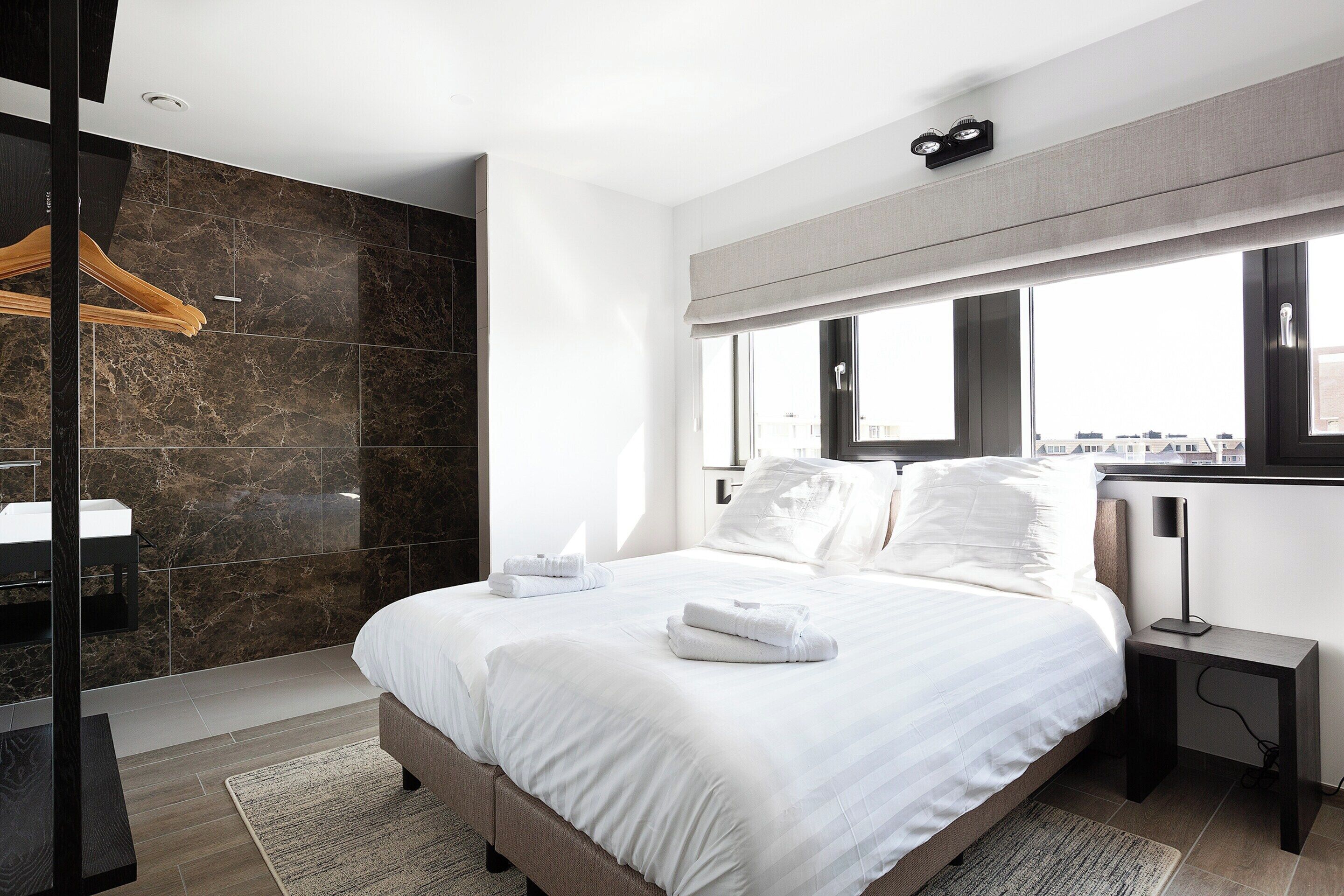 Quarto de hotel moderno com uma cama grande, mesa de cabeceira, espelho e vista da cidade.