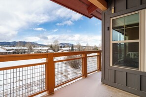 Ein Balkon mit Blick auf verschneite Landschaft und ferne Häuser, einem Holzgeländer und ein Fenster mit Ausblick auf Himmel und Berge.
