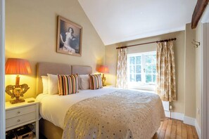 A bedroom with a bed, two bedside lamps, a window with curtains, a painting on the wall, and a wooden floor.