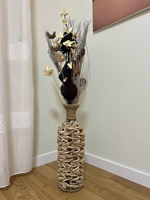 A wicker vase with a woven design, containing dried flowers and foliage, placed on a wooden floor against a white wall.