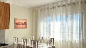 Ein Essbereich mit Tisch und Stühlen, einem Bild an der Wand, einem Fenster mit transparenten Vorhängen und einer Deckenleuchte.