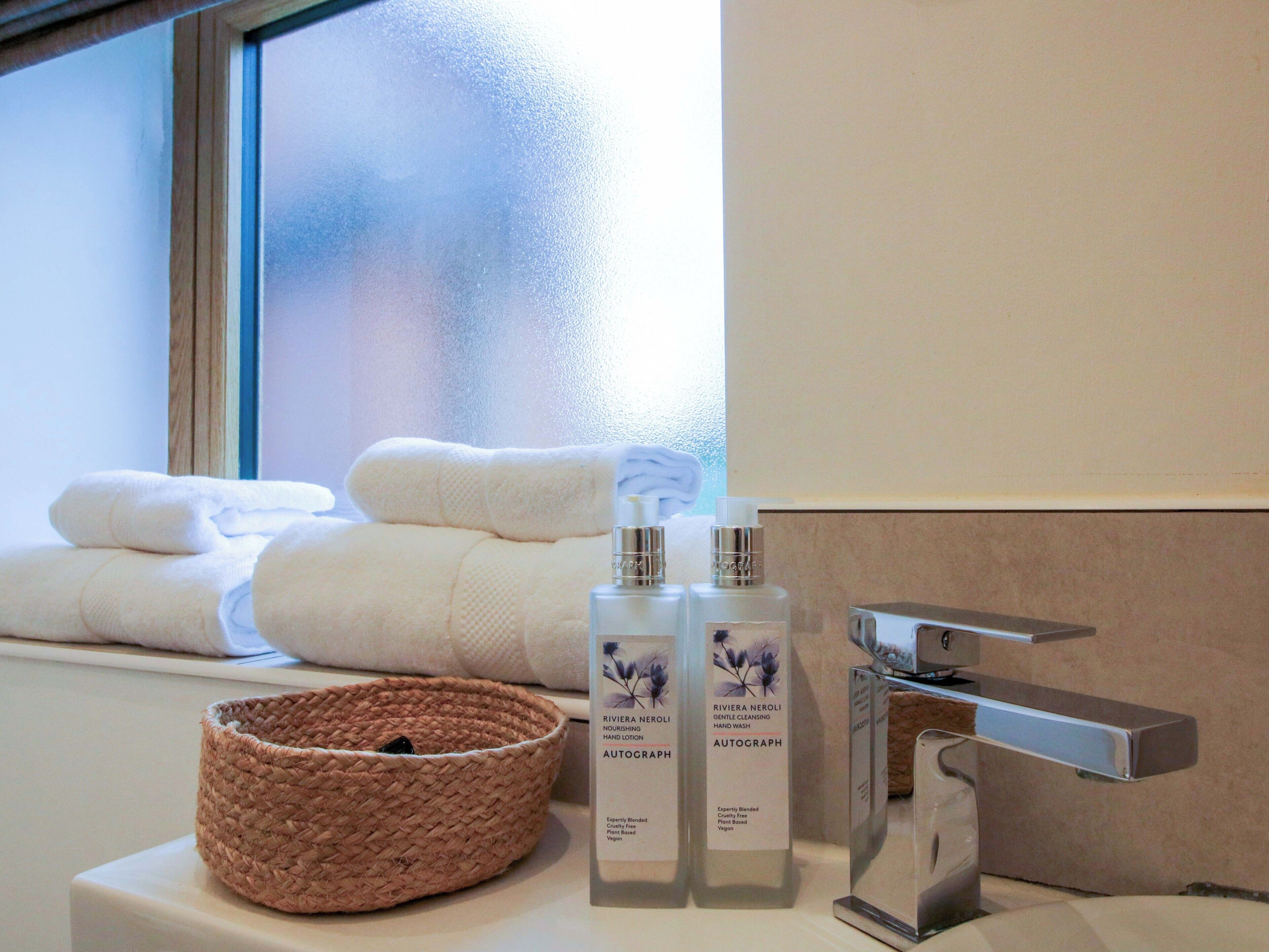 A bathroom with a window, two bottles of hand lotion, a woven basket, and a towel.
