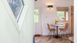 A dining area with a round table, two chairs, a wall-mounted light fixture, and a window with a view of greenery. - The Blacksmiths Shop (Bromyard)
