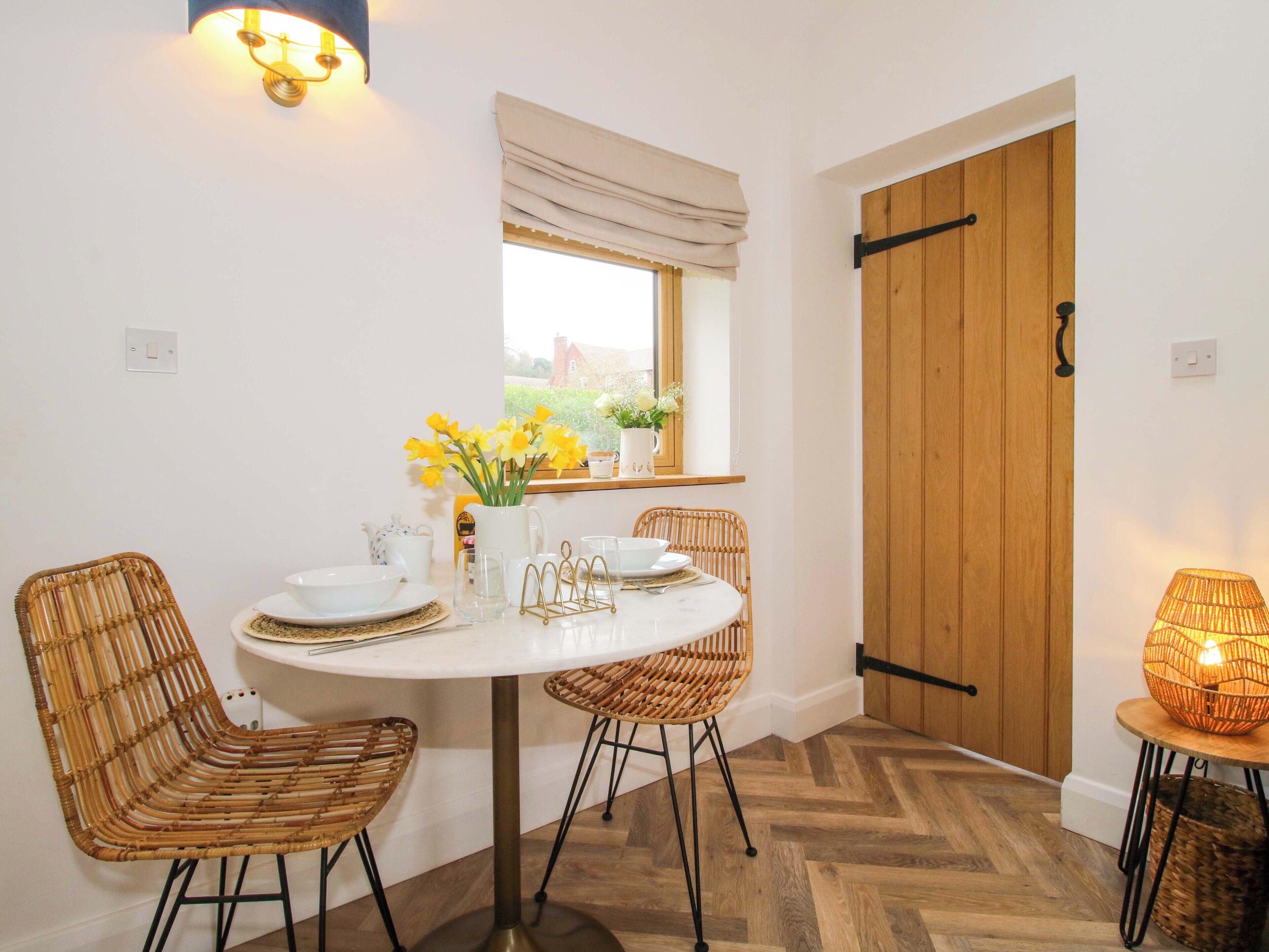 A dining area with a round table, four wicker chairs, a vase with flowers, and a wooden door.