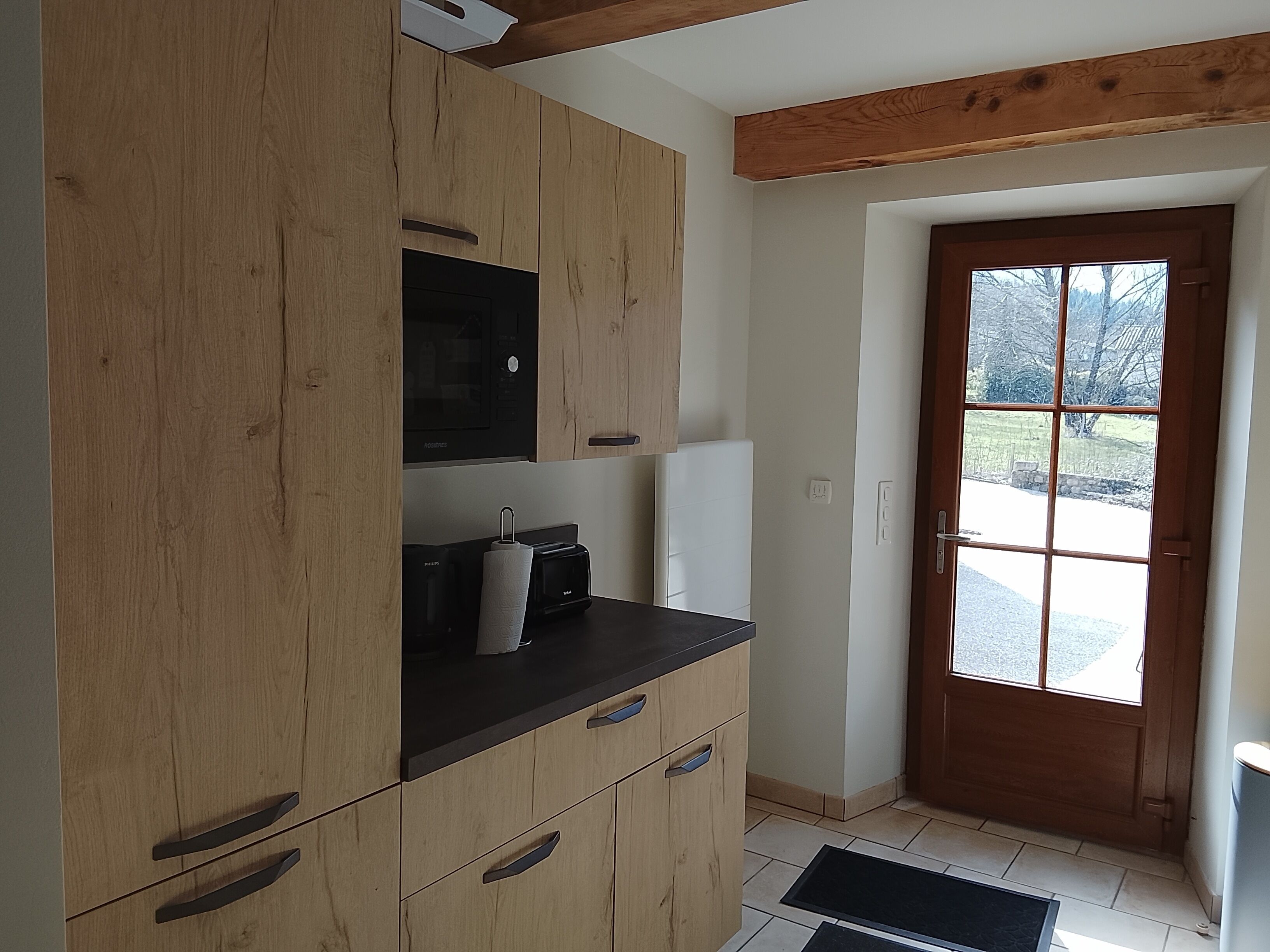 A kitchen with wooden cabinets, a black countertop, a microwave, and a door leading to a view of a snowy landscape.