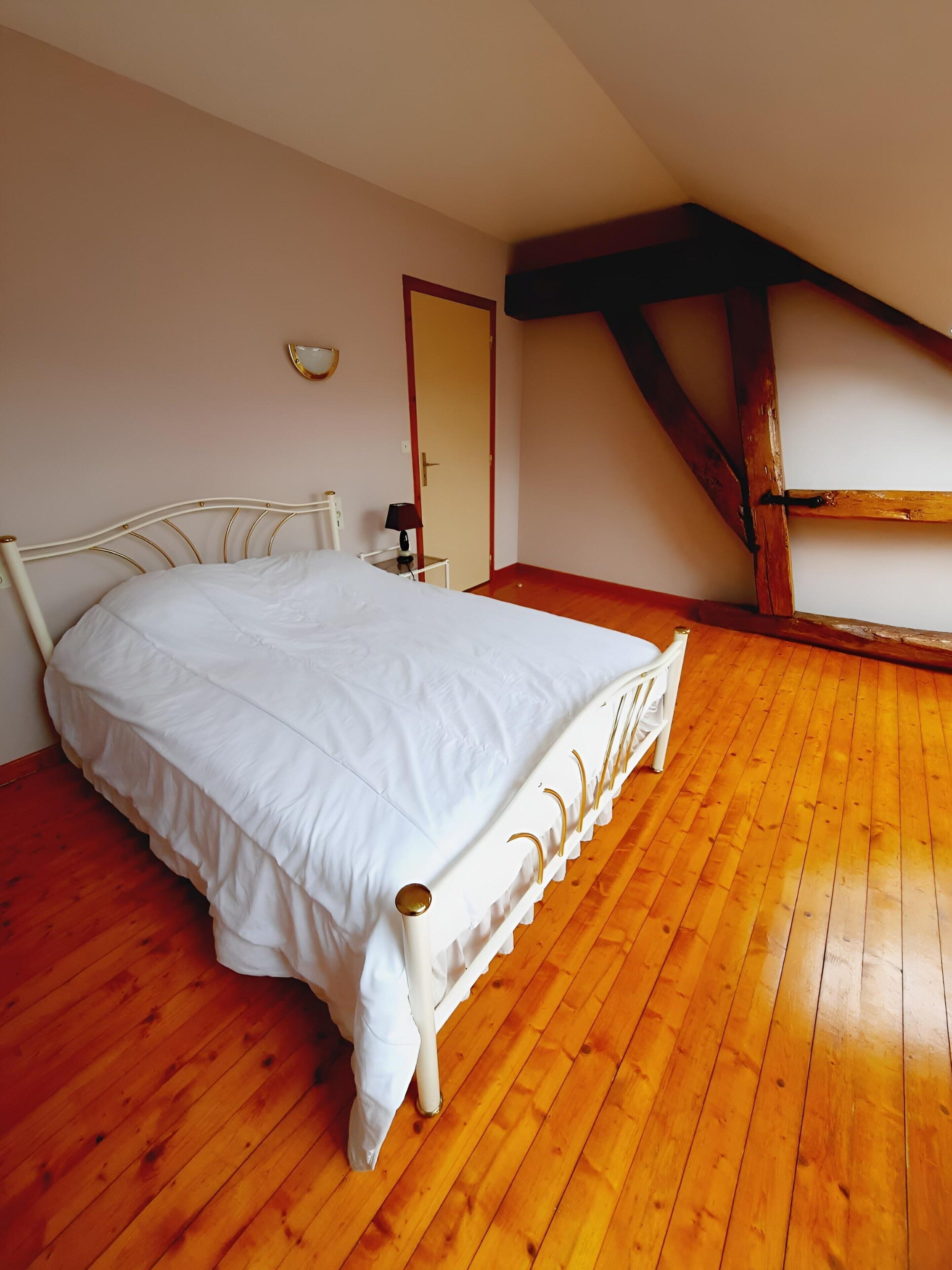 A bedroom with a white bed, wooden flooring, a small bedside table, and a door.