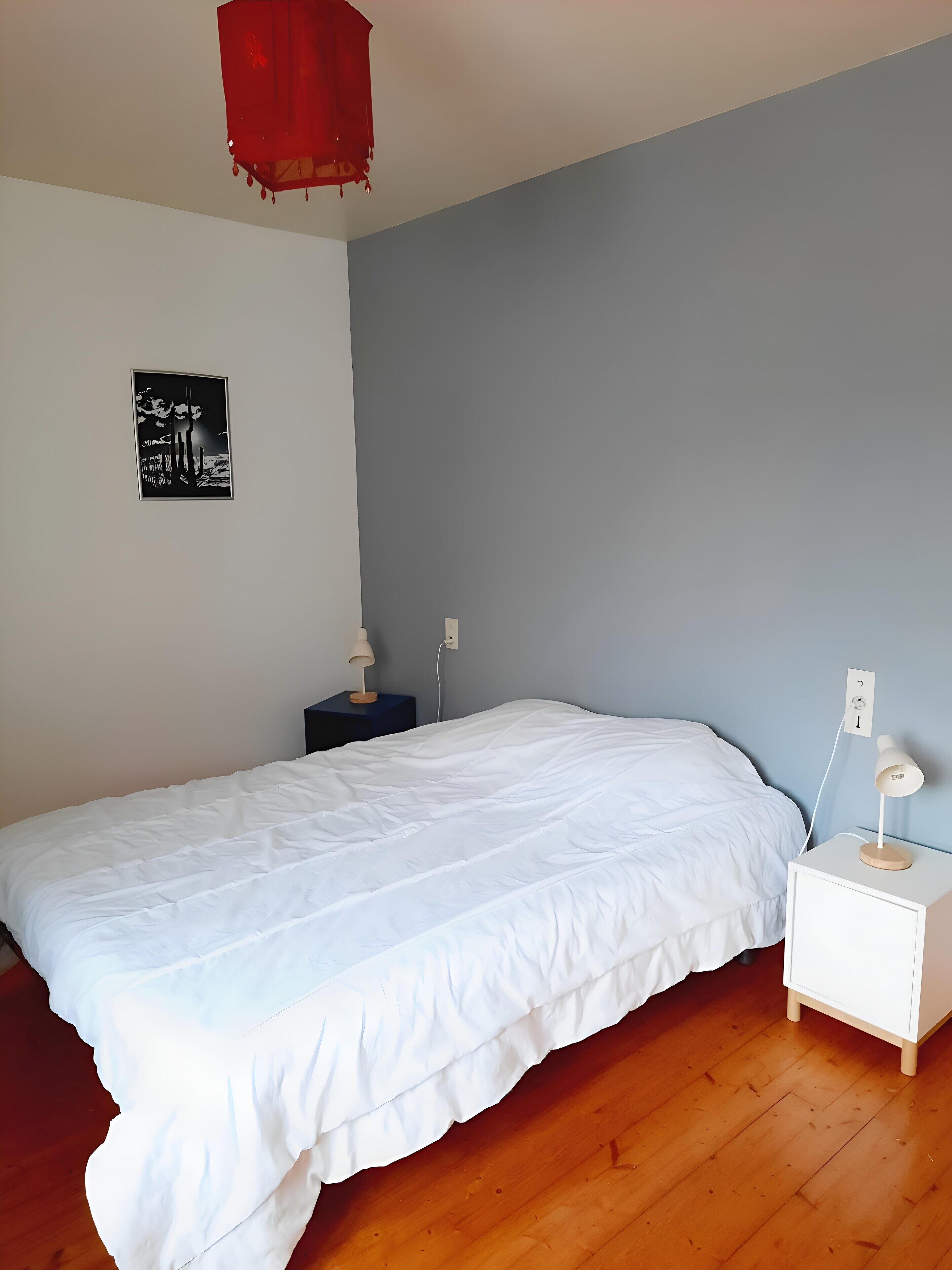 Une chambre à coucher comprenant un lit, une table de chevet, une lampe suspendue rouge et une œuvre d’art accrochée au mur.