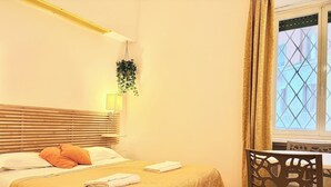 A hotel room with a wooden headboard, a bed with a patterned bedspread, a bedside table with a lamp, and a window with curtains. - Studio Apartment 4 pax (Roma)