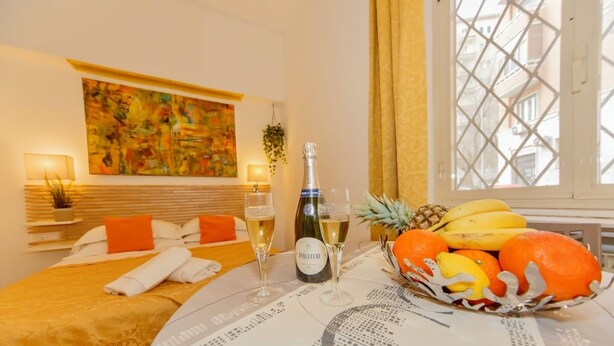A hotel room with a bed, a table with a bottle of champagne, glasses, and a fruit basket. - Studio Apartment 4 pax (Roma)