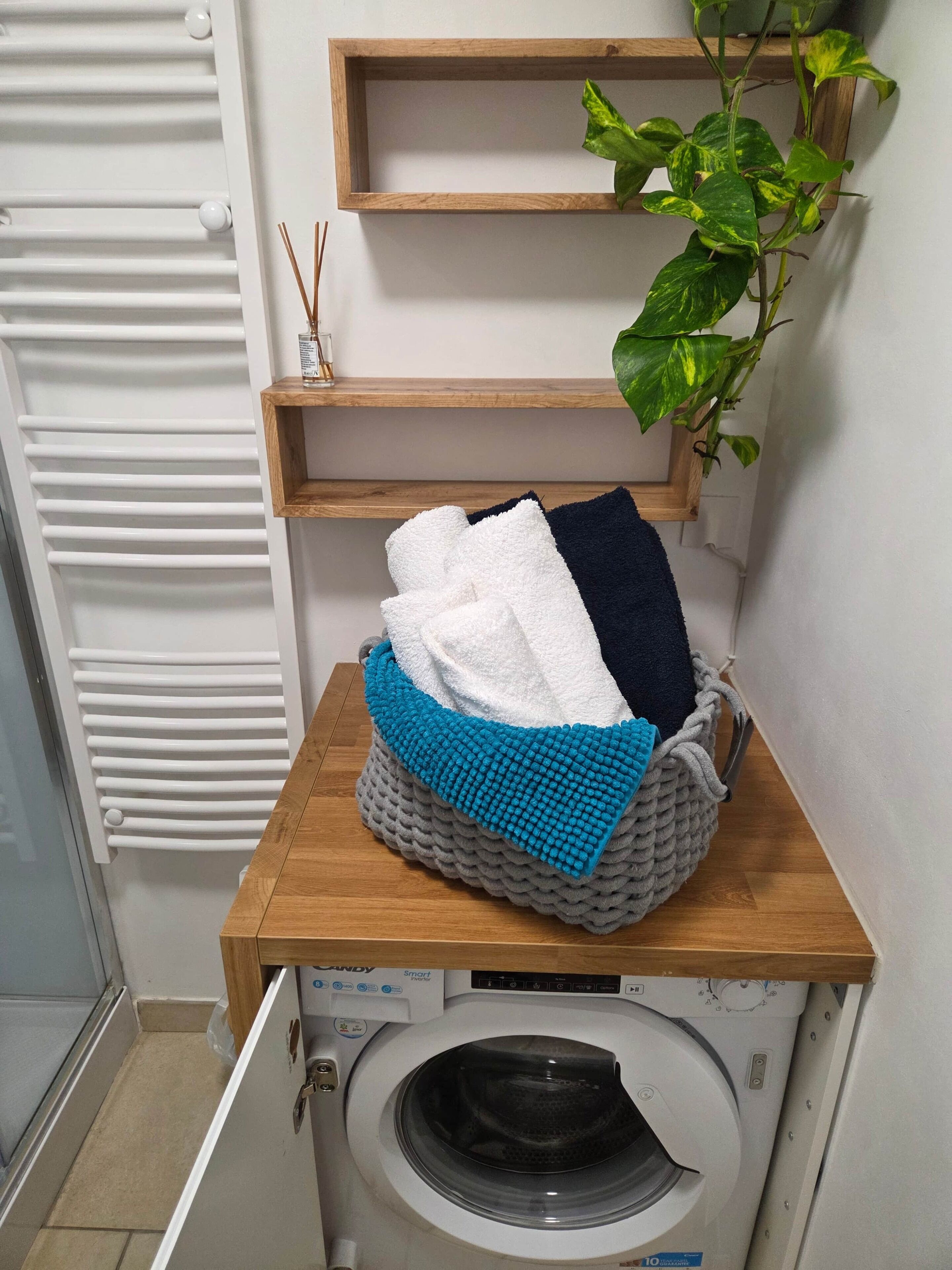 Un área de lavandería con lavadora, un estante de madera con una planta y una cesta de toallas dobladas.