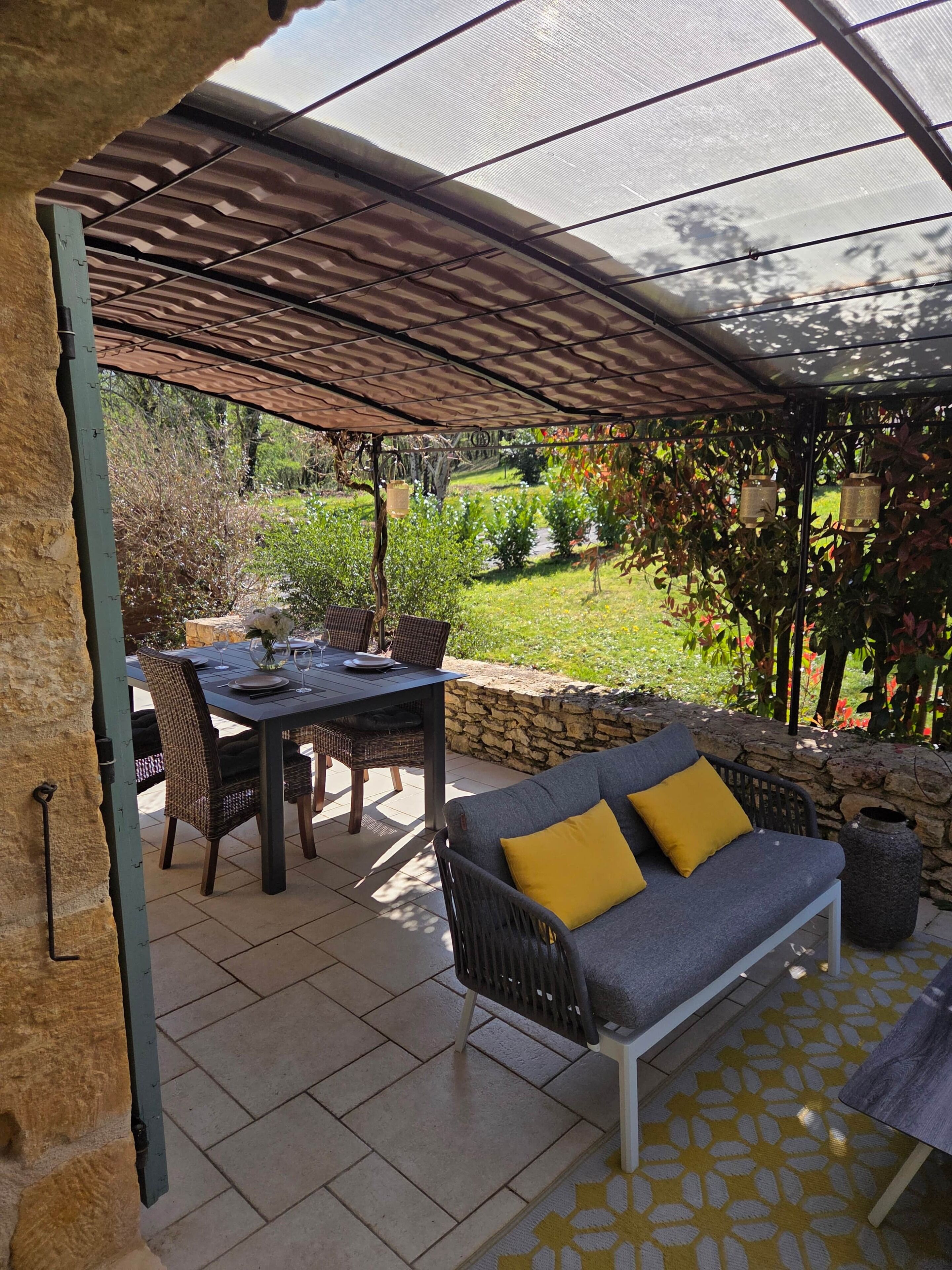 Un patio cubierto con una mesa, sillas, un sofá con cojines, un muro de piedra y vistas a un jardín.