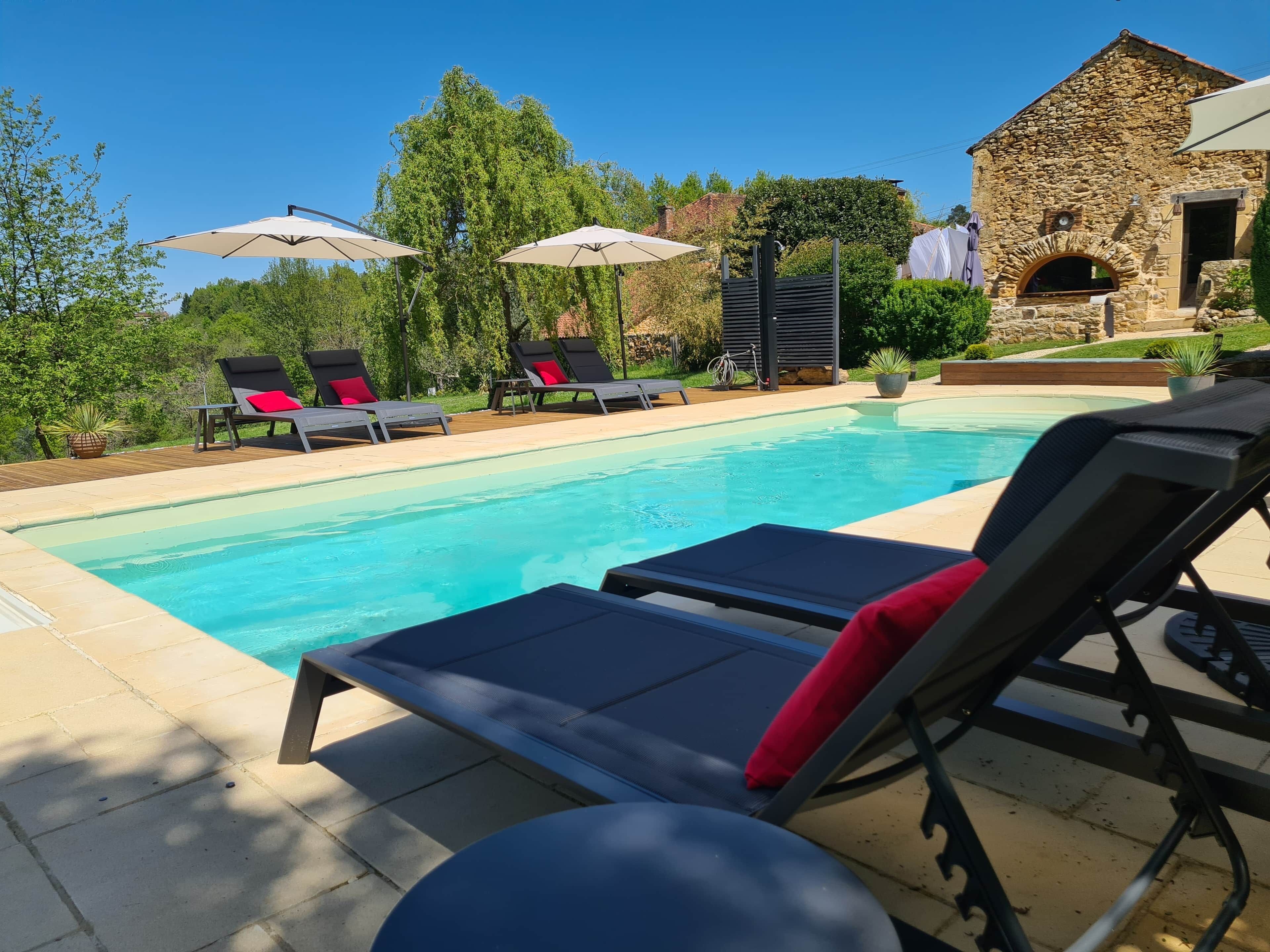 A swimming pool with lounge chairs and umbrellas, surrounded by greenery and a stone building.