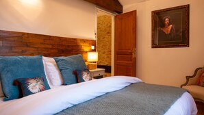 Quarto com cabeceira de madeira, cama com roupa de cama branca, mesa de cabeceira com abajur, porta e quadro na parede.