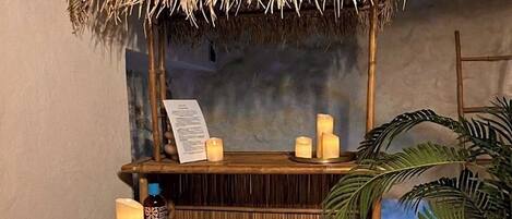 A room with a bed, a small wooden shelf with candles, a bottle, and a towel.