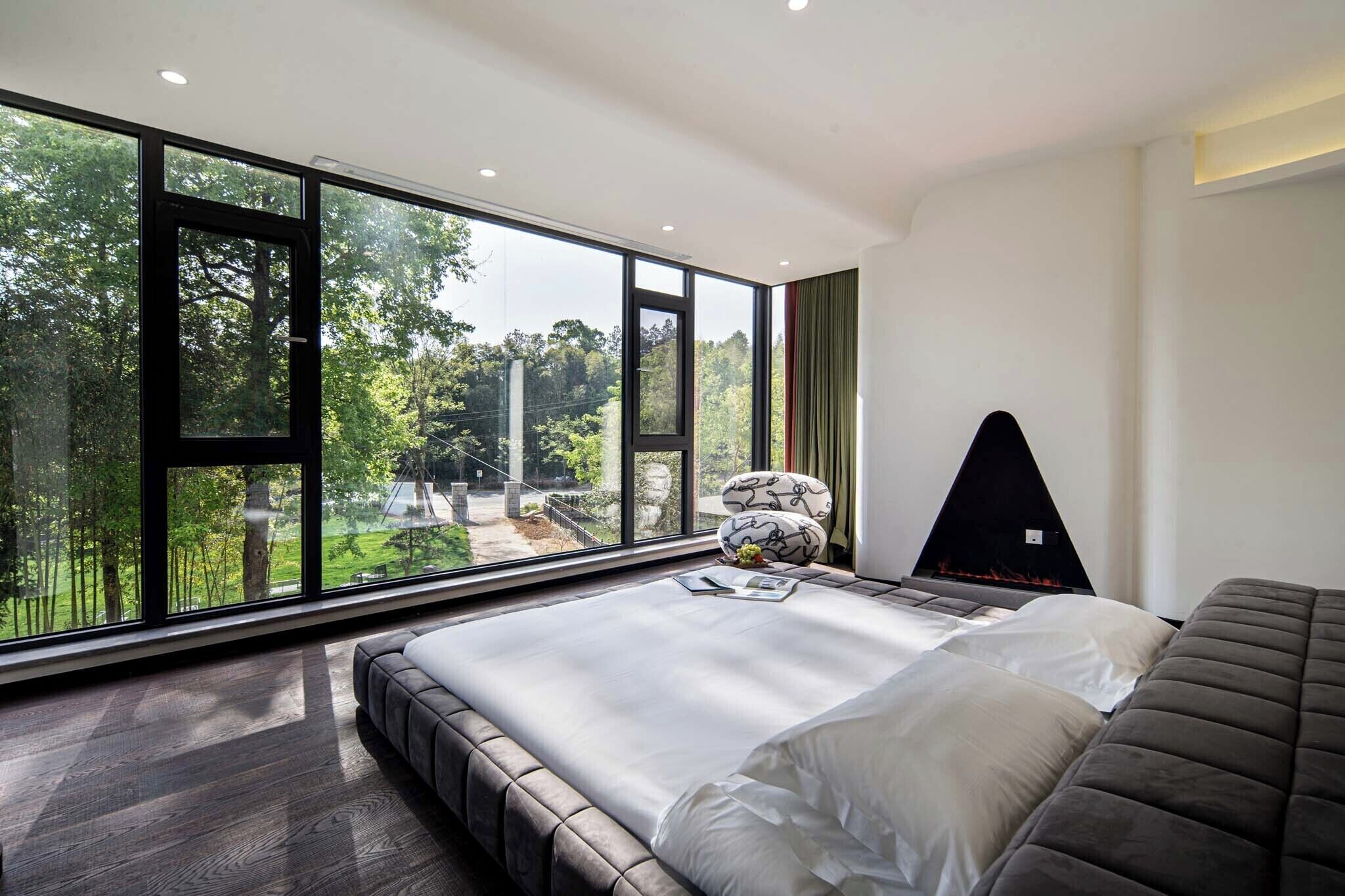 A modern bedroom with a large window, a bed, a seating area, and a fireplace.