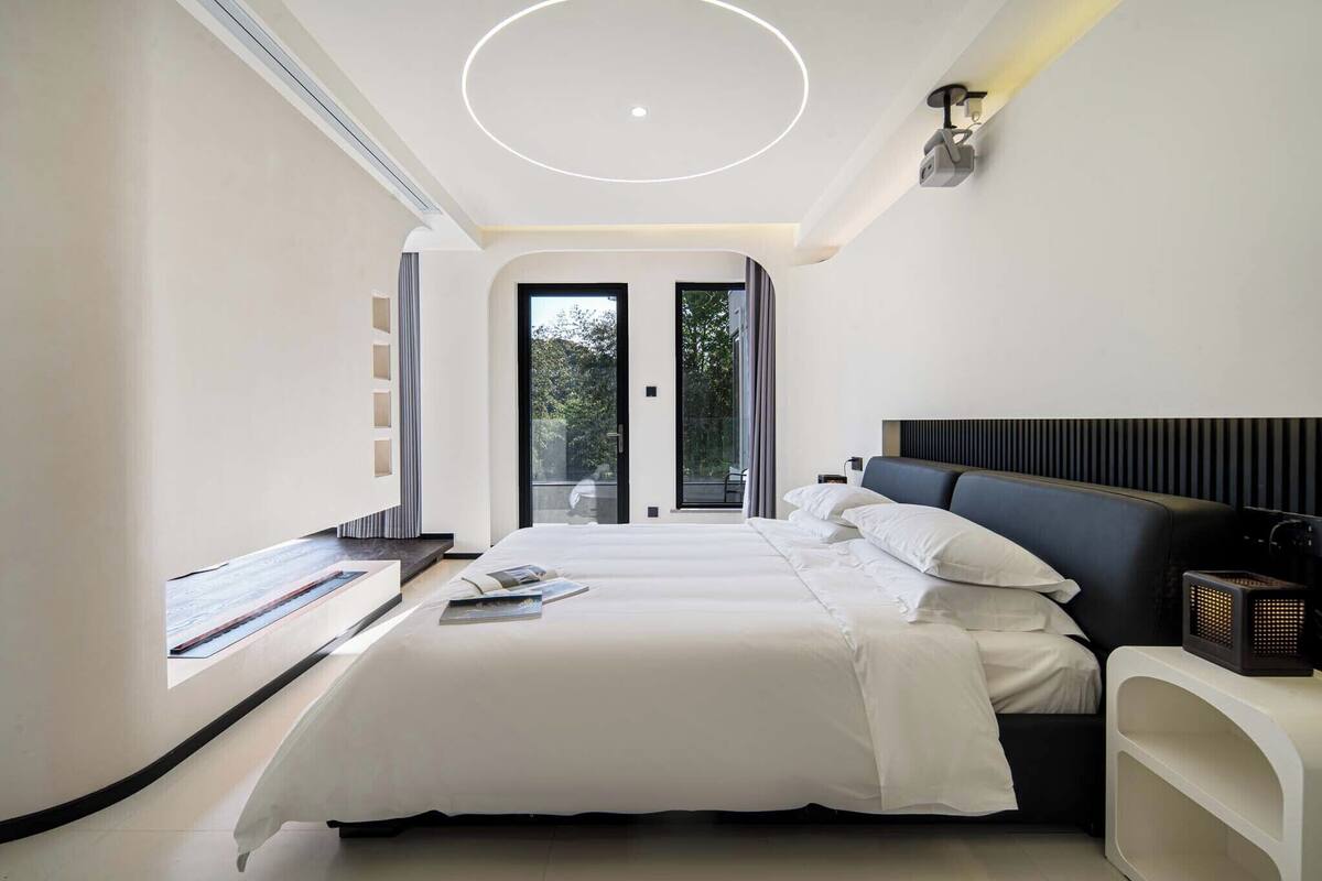 Une chambre d’hôtel moderne dotée d’un grand lit, d’une table de chevet, d’un coin lecture avec fenêtre et d’un luminaire au plafond.