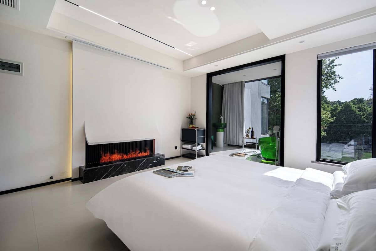 Une chambre moderne avec une cheminée, un lit, un fauteuil vert et une vue sur l’extérieur grâce à de grandes fenêtres.