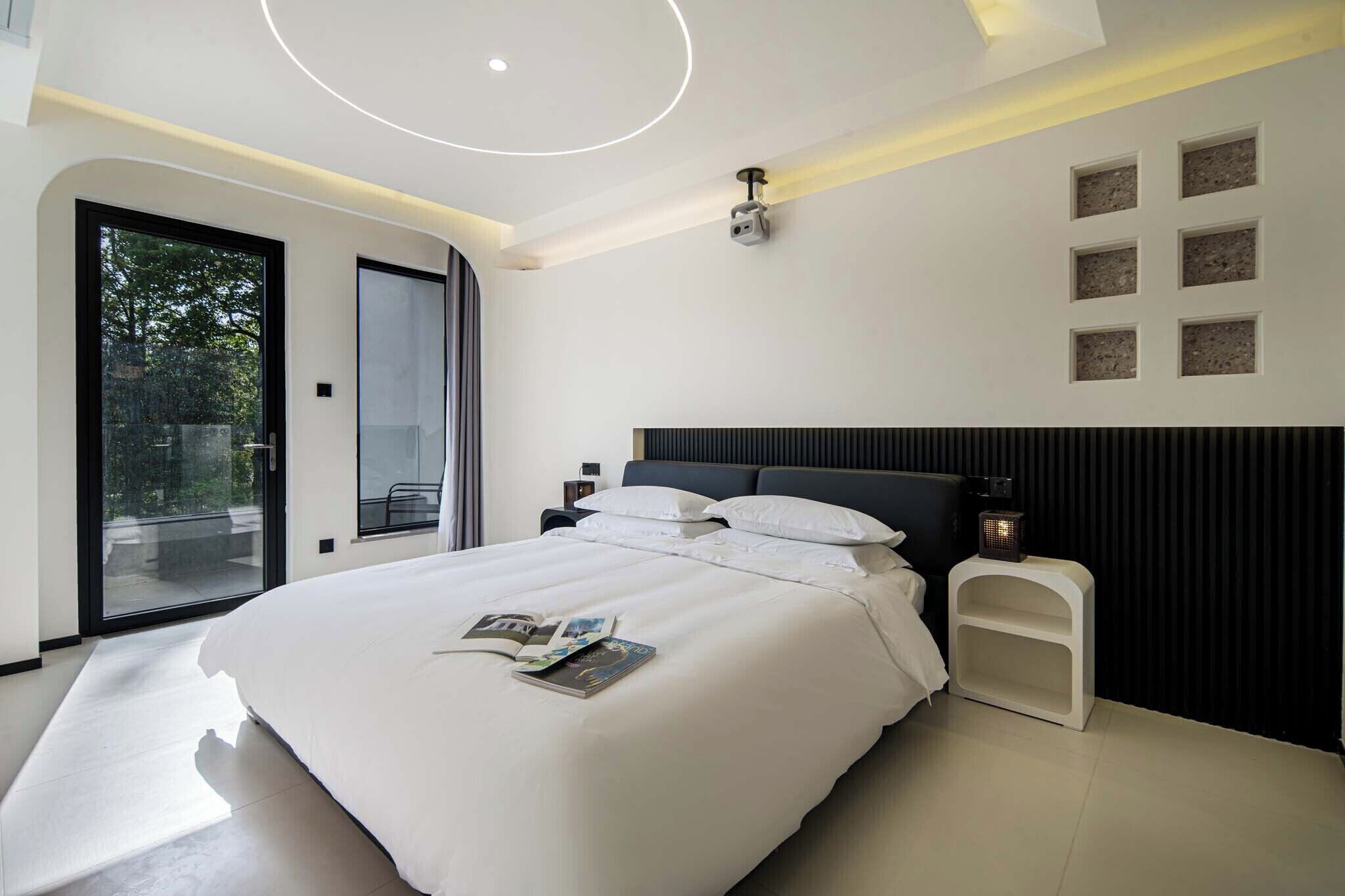 Une chambre moderne avec un grand lit, une porte-fenêtre coulissante donnant sur l’extérieur, un luminaire au plafond et une petite table avec une lampe.