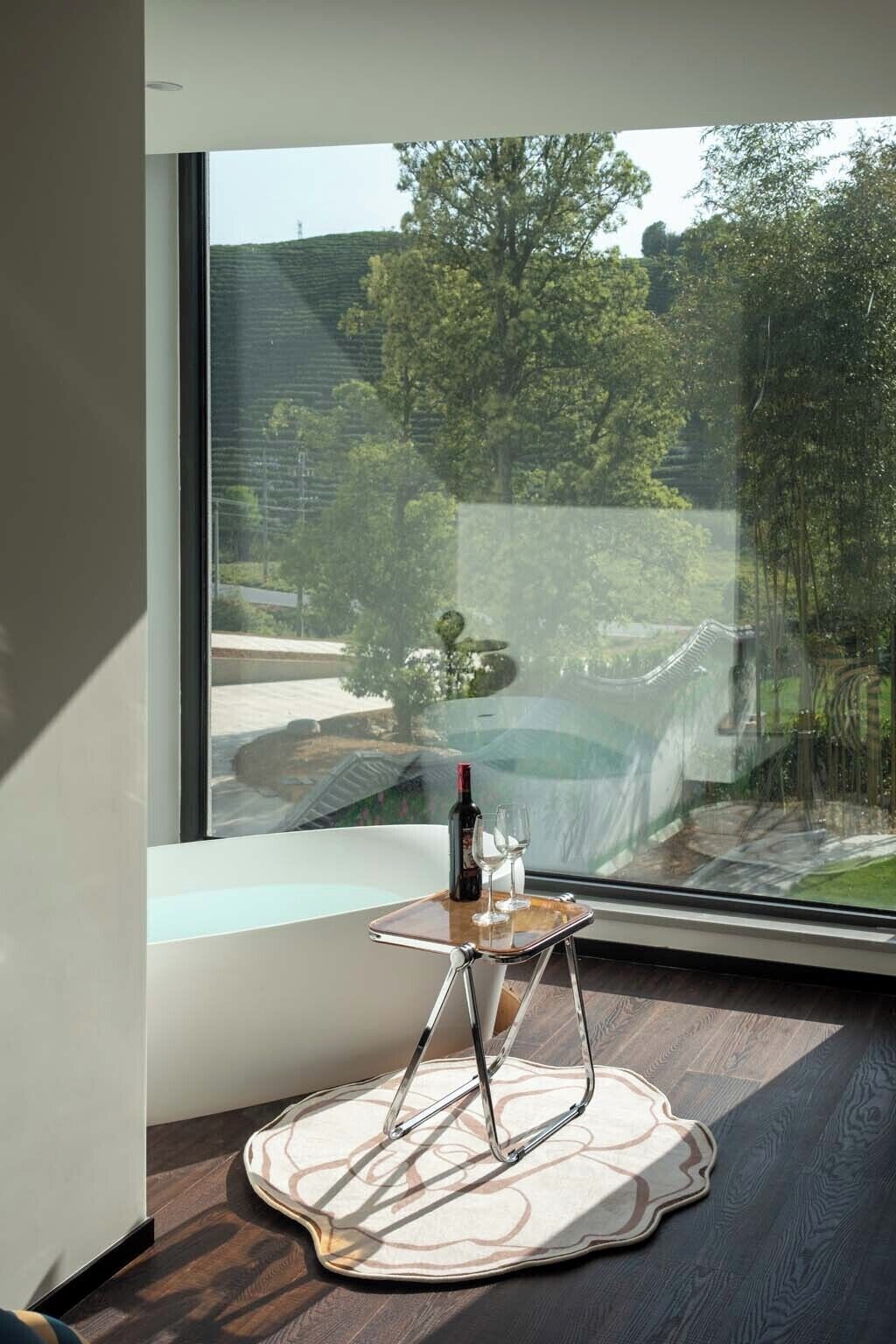 A modern room with a large window, a small table with a bottle and glass, and a patterned rug.