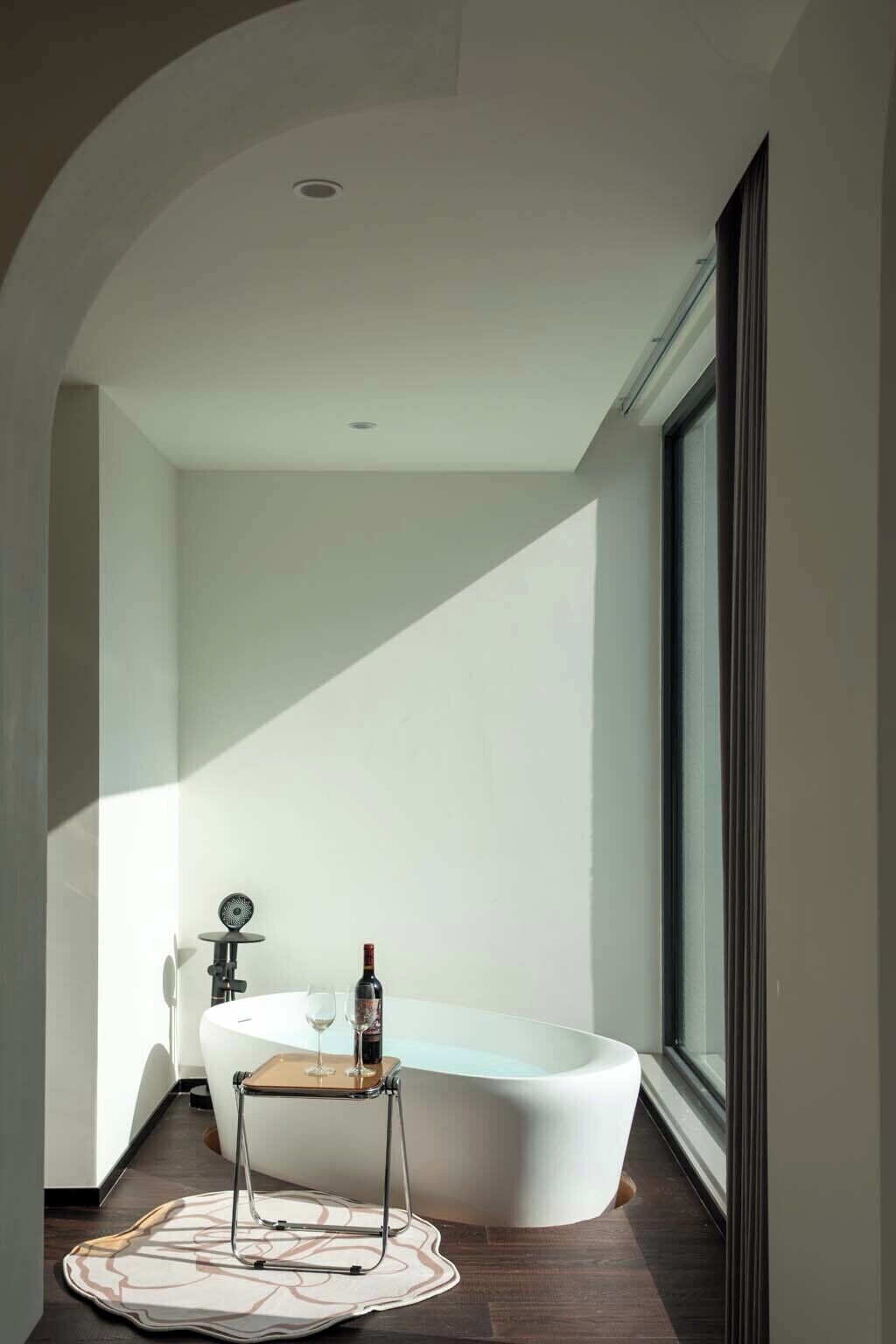 A modern bathroom with a freestanding bathtub, a small table with a wine bottle, and a large window providing natural light.