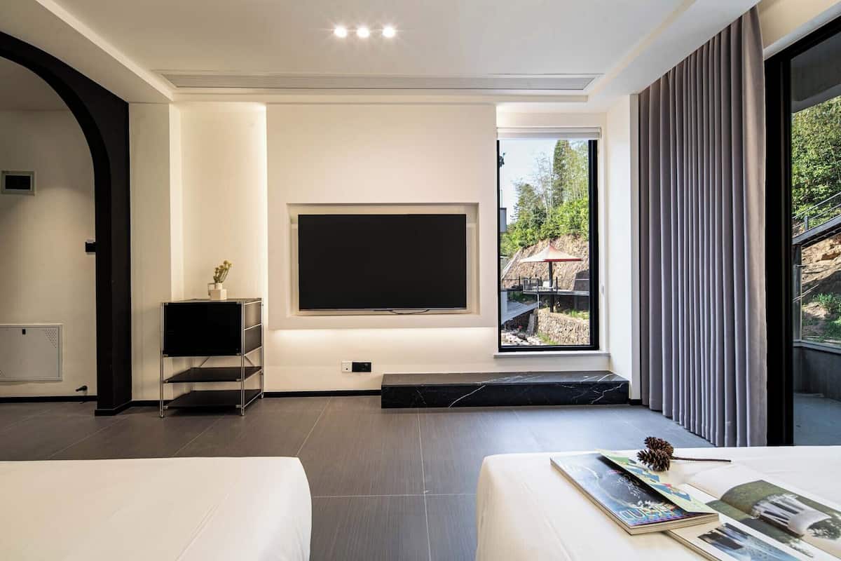 Une chambre d’hôtel moderne, dotée d’une grande télévision, d’un lit, d’une petite table avec un livre et d’une fenêtre avec des rideaux.
