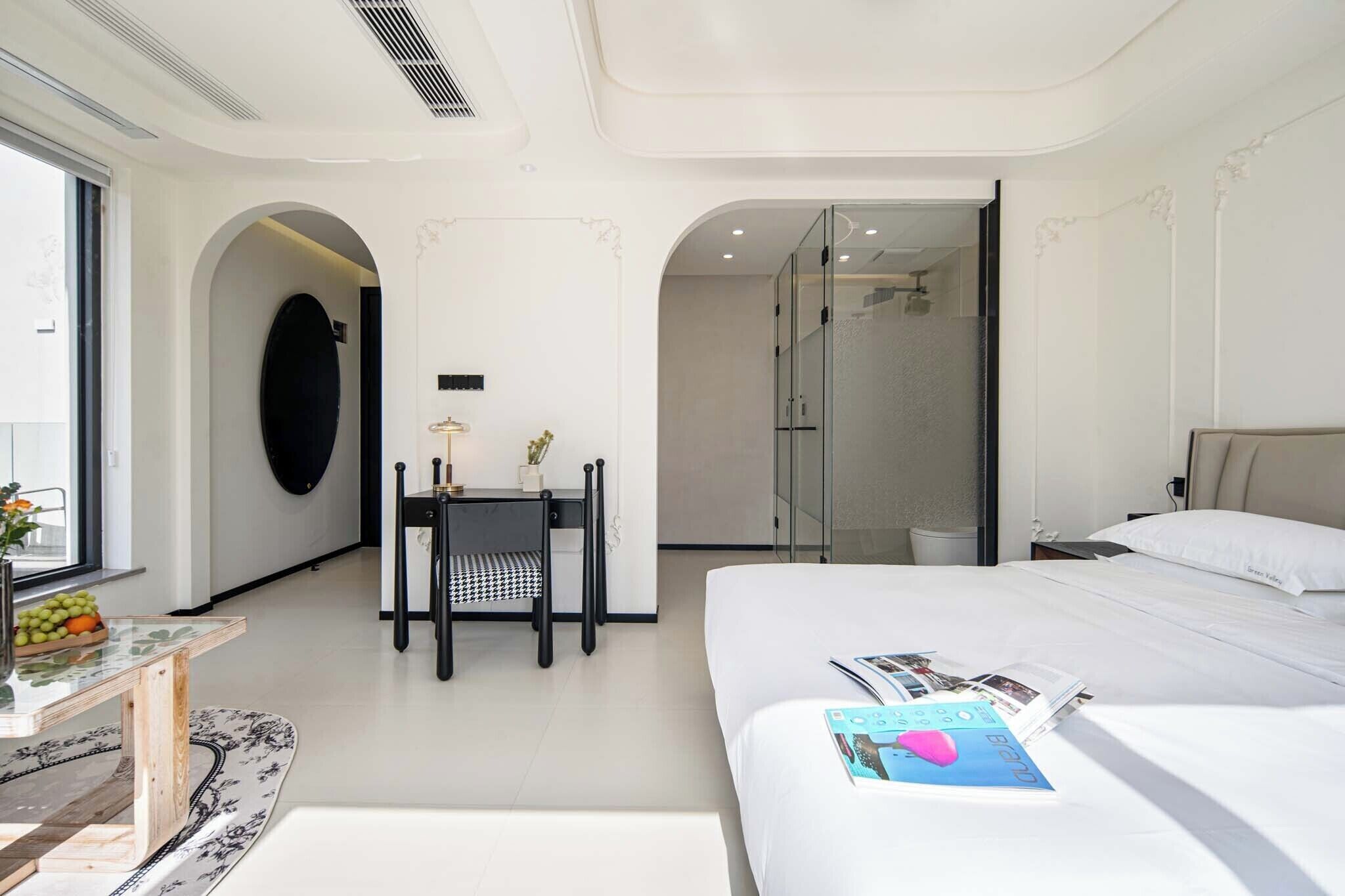 Ein Hotelzimmer mit einem Bett, einem Schreibtisch mit Stuhl, einem kleinen Tisch mit Vase und Früchten sowie einer separaten Duschkabine.