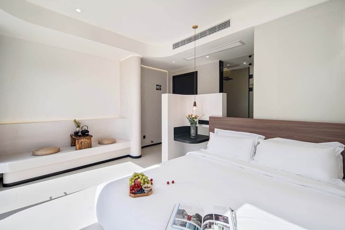 Une chambre d’hôtel moderne avec un grand lit, une table de chevet agrémentée d’un vase, un petit coin salon avec des coussins et une étagère fixée au mur.