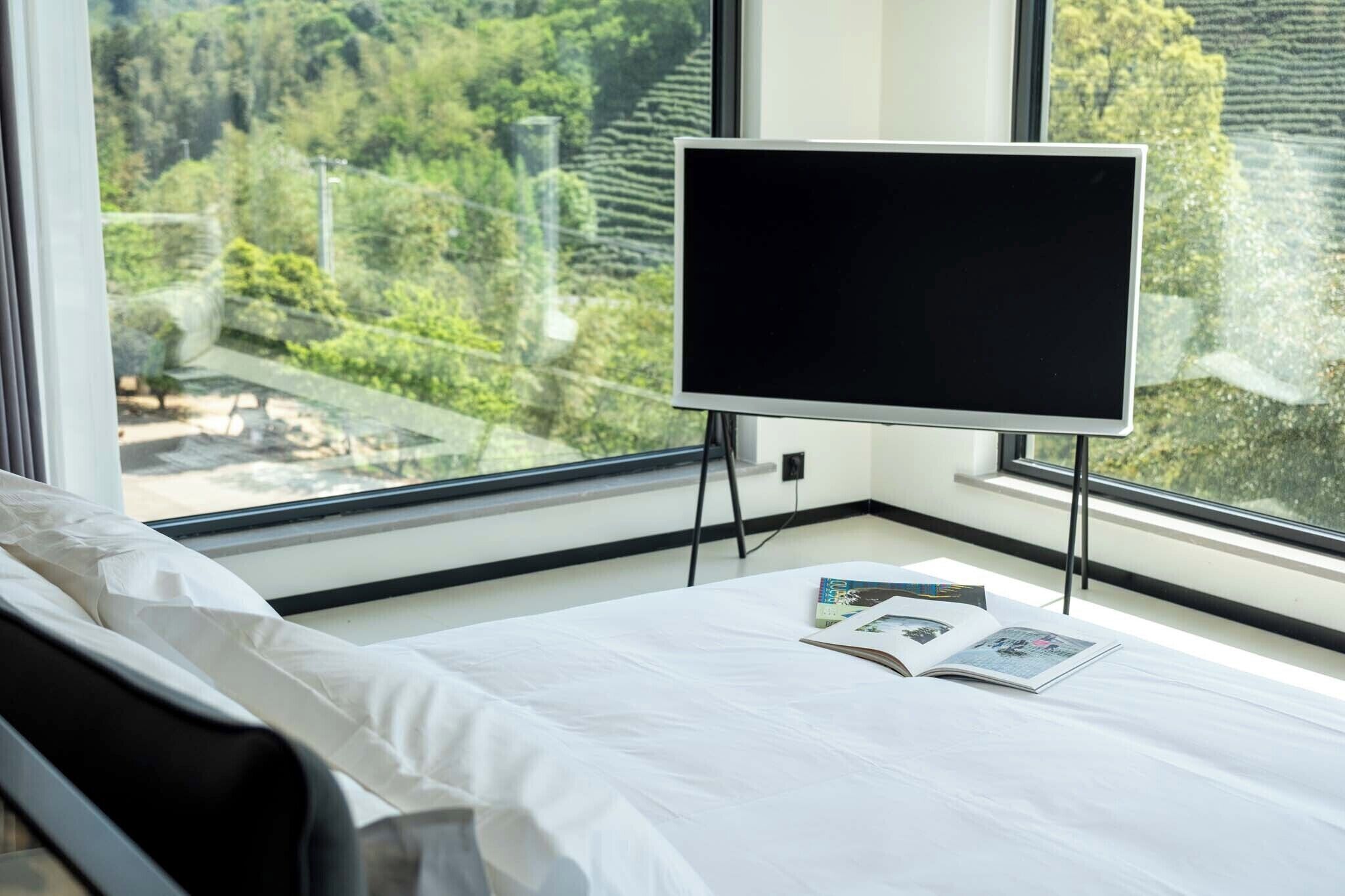 Ett hotellrum med en säng, en platt-TV och utsikt över en skog genom stora fönster.