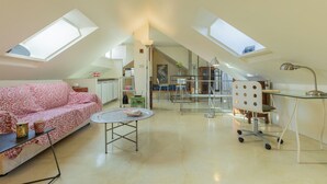Um loft espaçoso com pé direito alto, claraboias, um sofá, uma mesa redonda e uma escrivaninha com abajur.