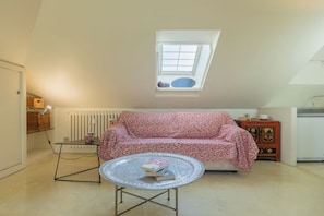 A cozy room with a sloped ceiling, a red patterned sofa, a small round table with books, a radiator, and a window with a view.