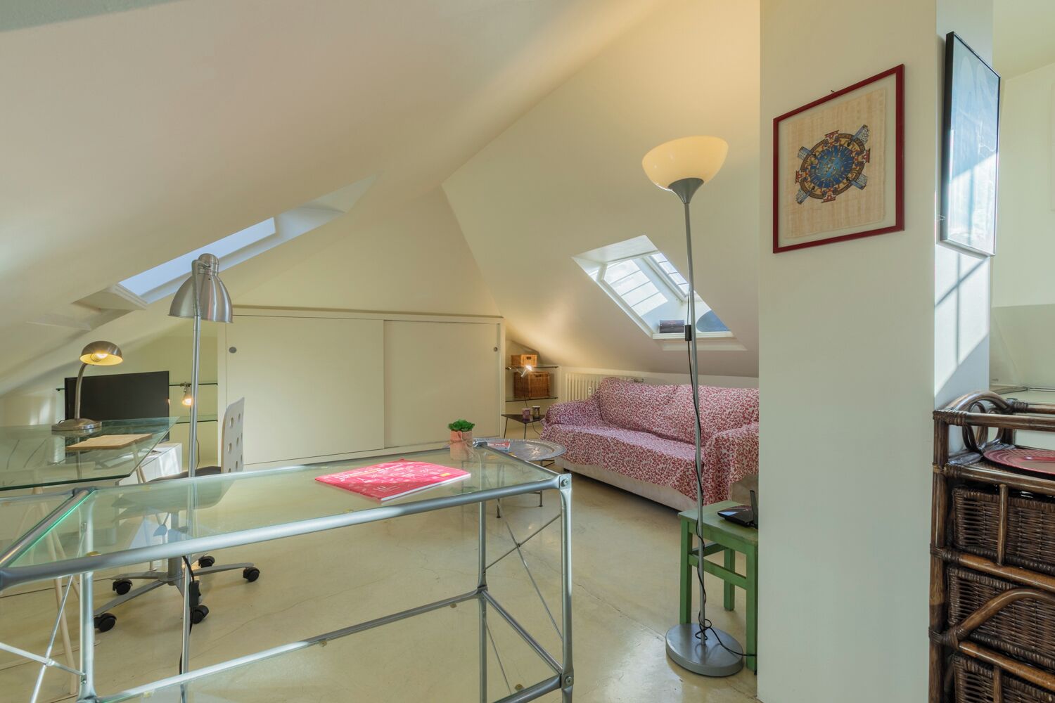 A loft-style room with a glass desk, a green stool, a red sofa, a floor lamp, and a framed picture on the wall.