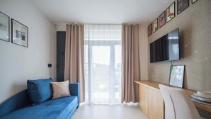 Ett modernt vardagsrum med en blå soffa, ett skrivbord i trä, en tv och ett stort fönster med gardiner.