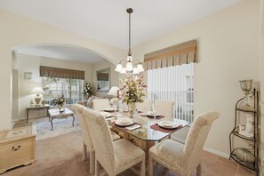 Un coin repas avec une table en verre, des chaises recouvertes d’un tissu beige, un lustre et un vase rempli de fleurs.
