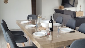 Una sala da pranzo moderna con un lungo tavolo apparecchiato per sei persone, circondato da sei sedie, una televisione appesa alla parete e un divano sullo sfondo.